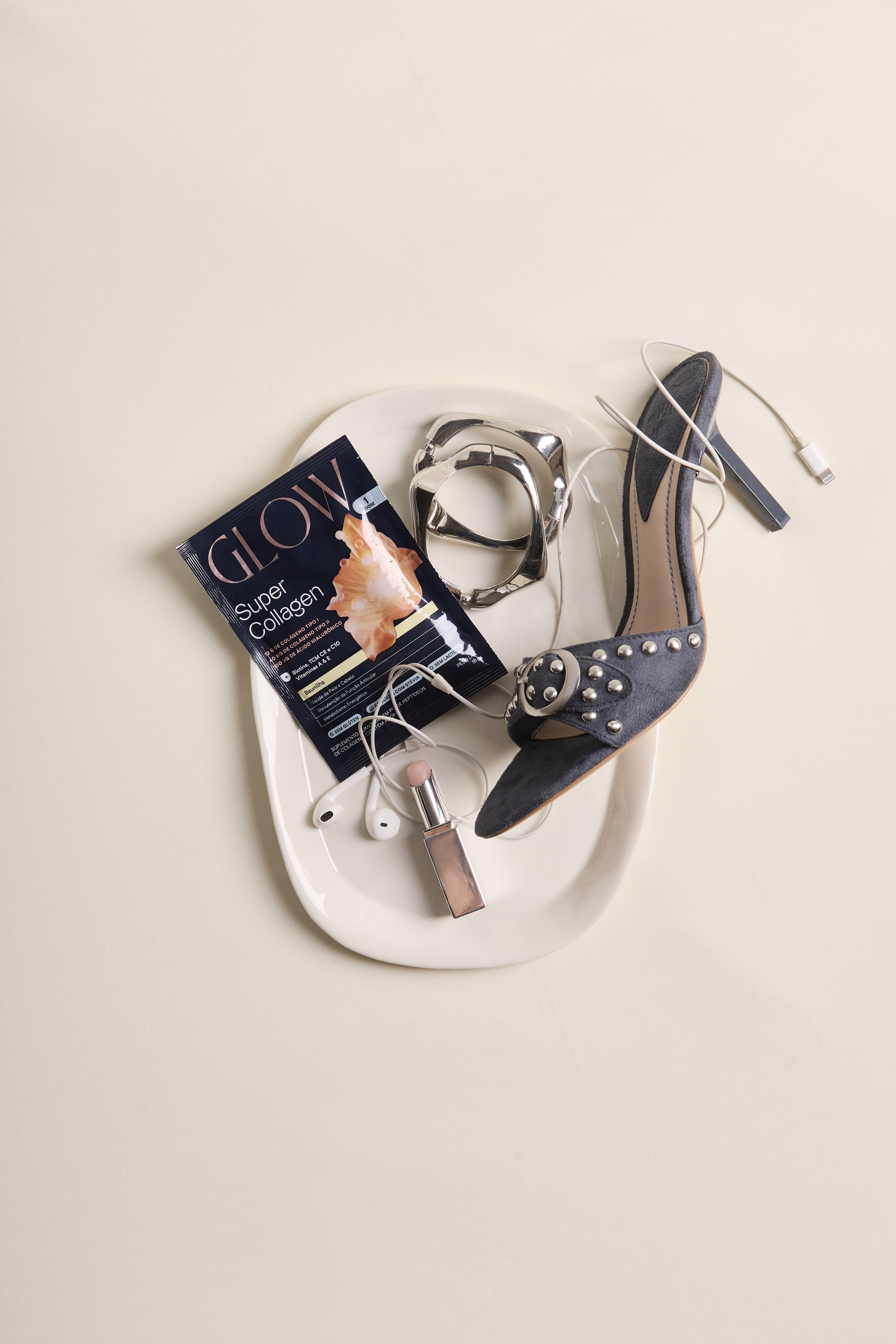 Embalagem sachê do suplemento Glow Super Collagen sobre uma bandeja branca, acompanhada de um salto alto azul com tachas, pulseira prateada, fones de ouvido brancos e um batom nude